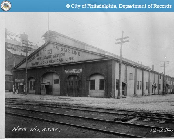 A large, two story building with the text "53 Immigrant Station, American Line Red-Star Line, Harrisburg-American Line" on the front. Railroad tracks are in front of the building and electric lines surround the top of the building. 